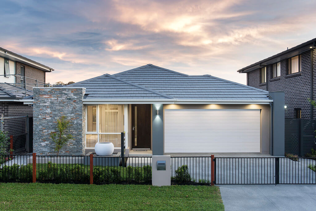 Modern single-story house with a gray tile roof, large white garage door, stone accent wall, fenced front yard, and a small lawn at sunset.